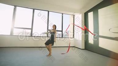 布鲁内特年轻女子舞蹈艺术体操，现代用红色丝带穿黑色连衣裙在窗户前。 太阳